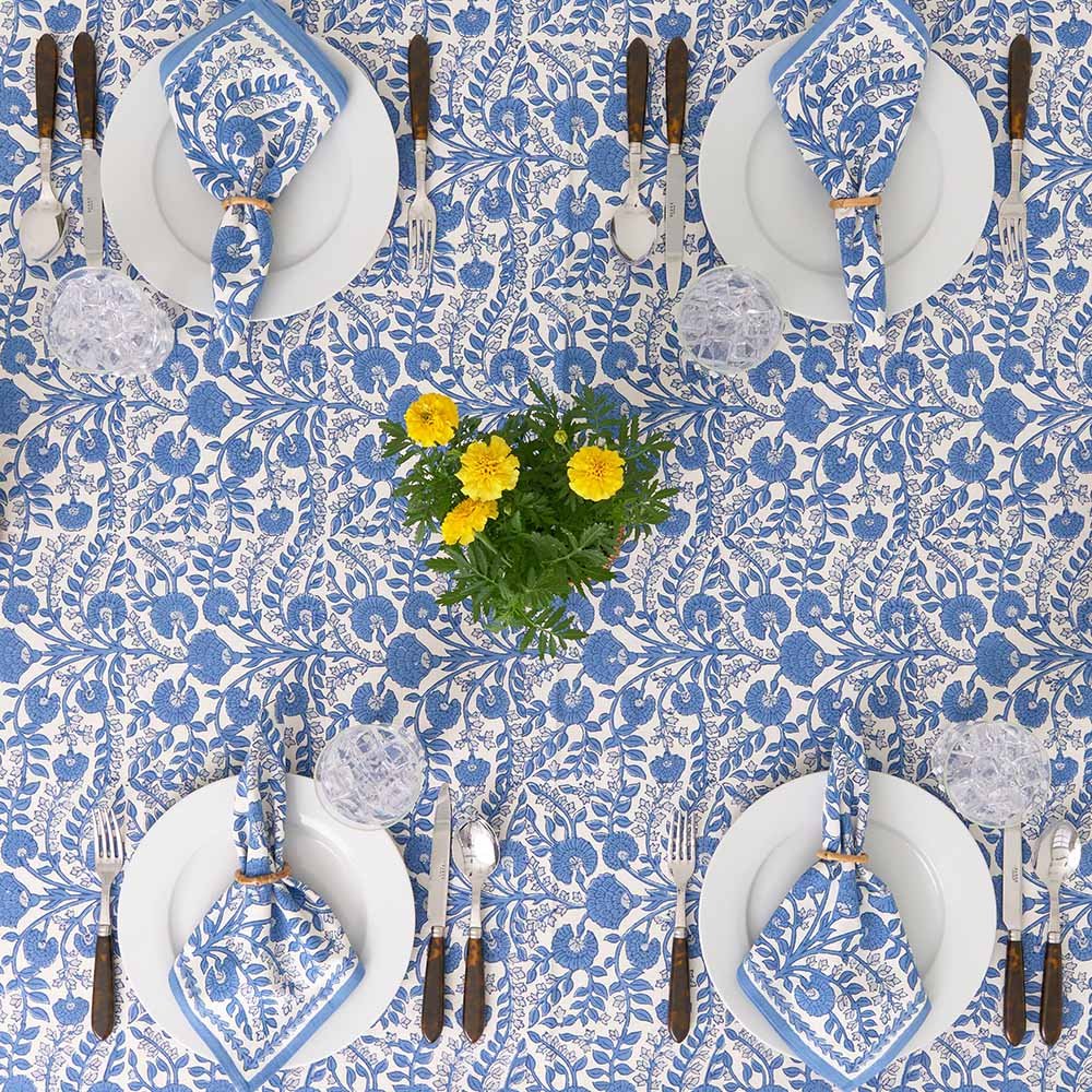 Table setting with blue floral tablecloth, white plates, silverware, and yellow flowers.