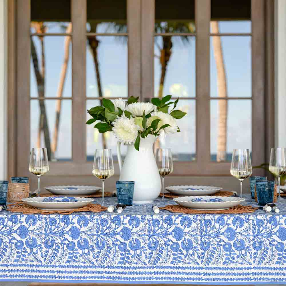 Elegant table setting with a floral tablecloth, plates, glasses, and a vase of flowers in a room with large windows.