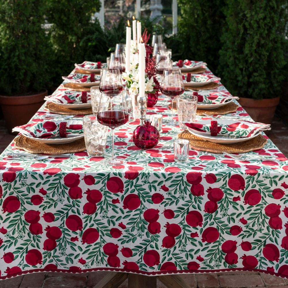 A tablecloth with a red pomegranate and green leaf pattern laid out on a table set for a meal, featuring dishes and glasses.