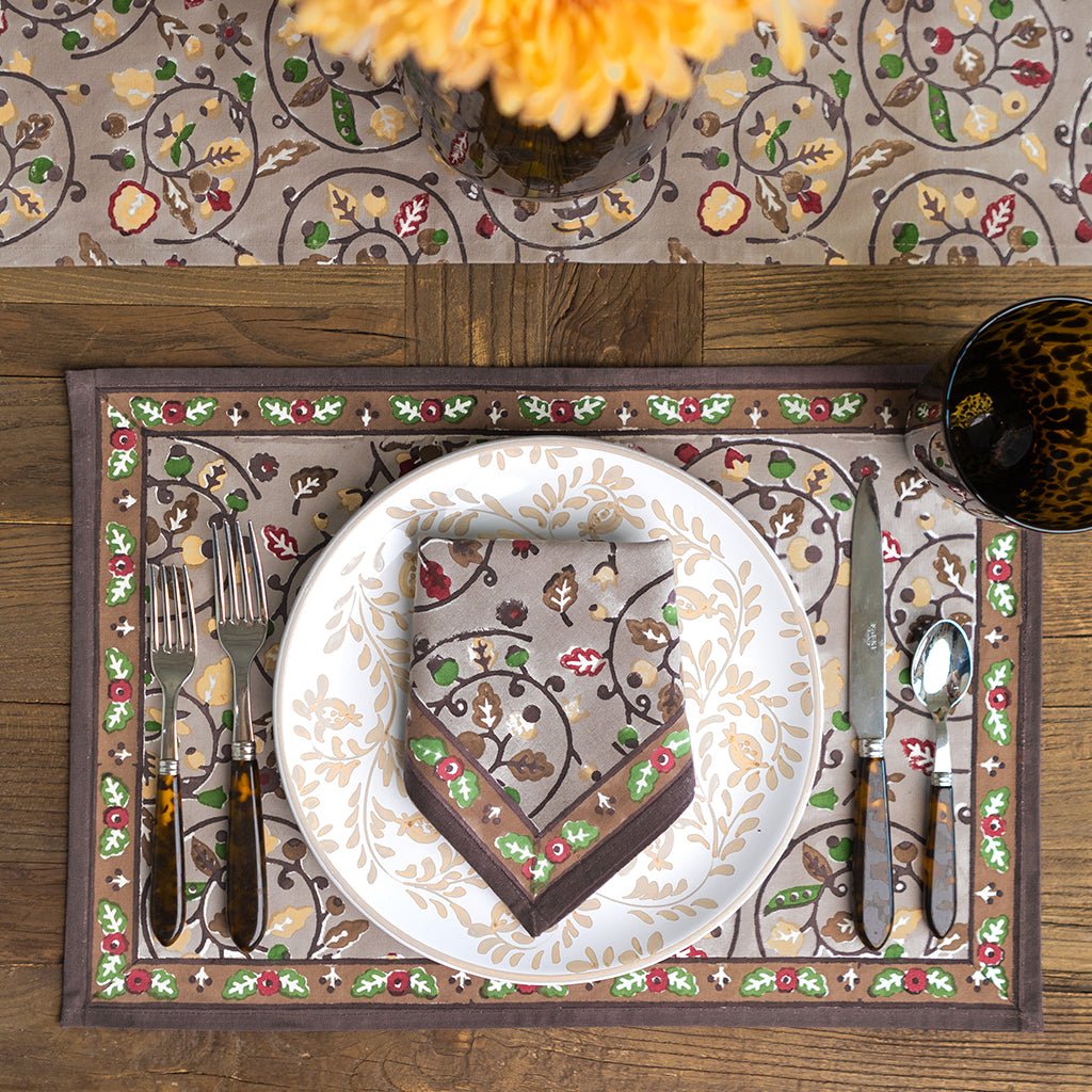 Decorative table setting with a brown patterned napkin on a plate, surrounded by cutlery and a floral-patterned placemat.