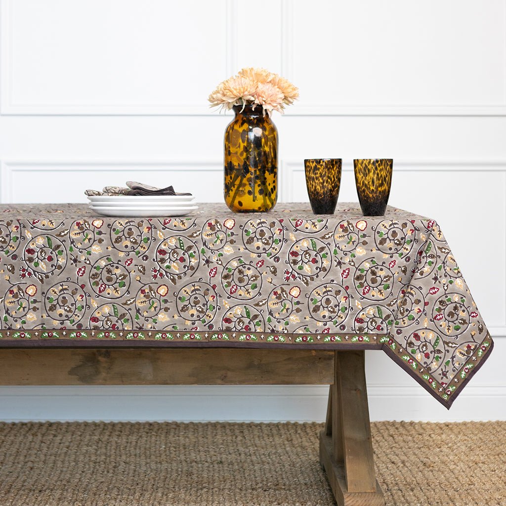 Tablecloth with floral pattern on a wooden table, with a vase and glasses in the background.