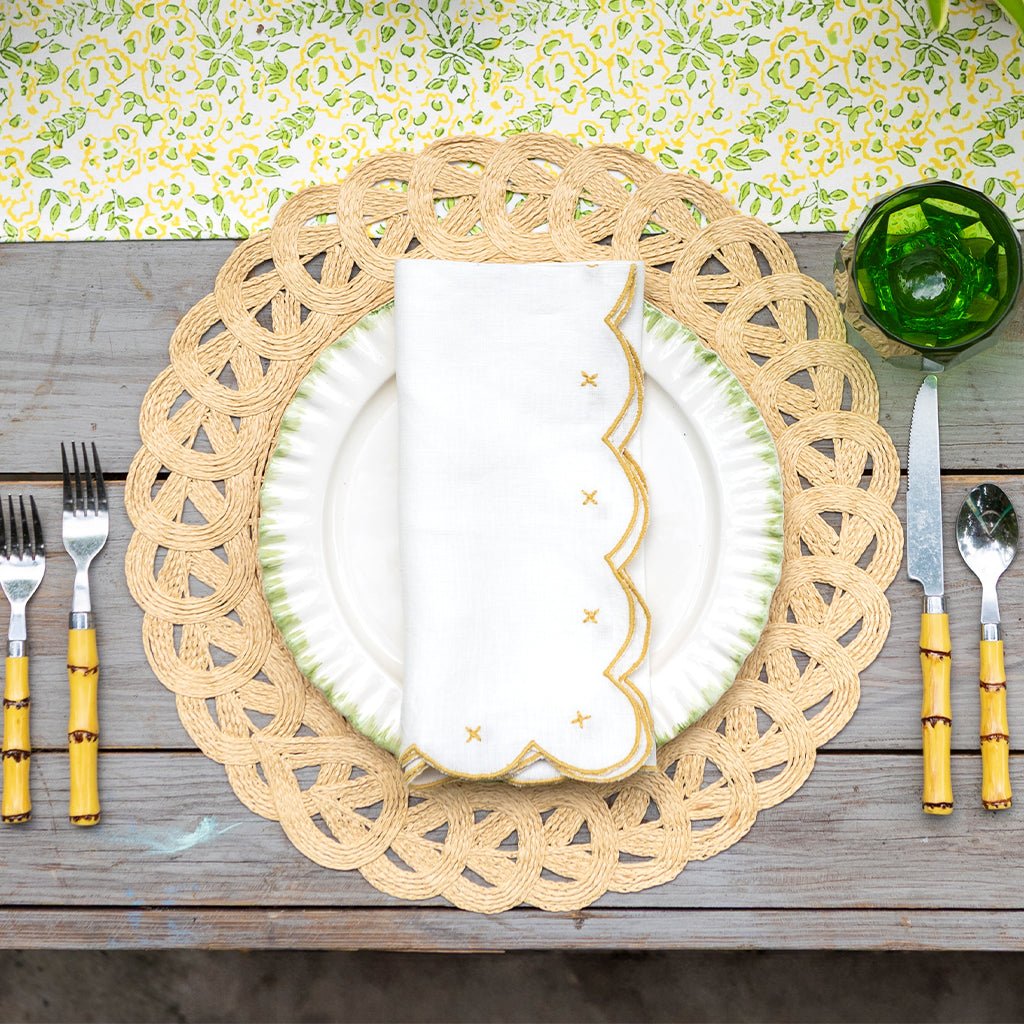 Table setting with a woven placemat, white plate with gold design, and cutlery on a wooden table.