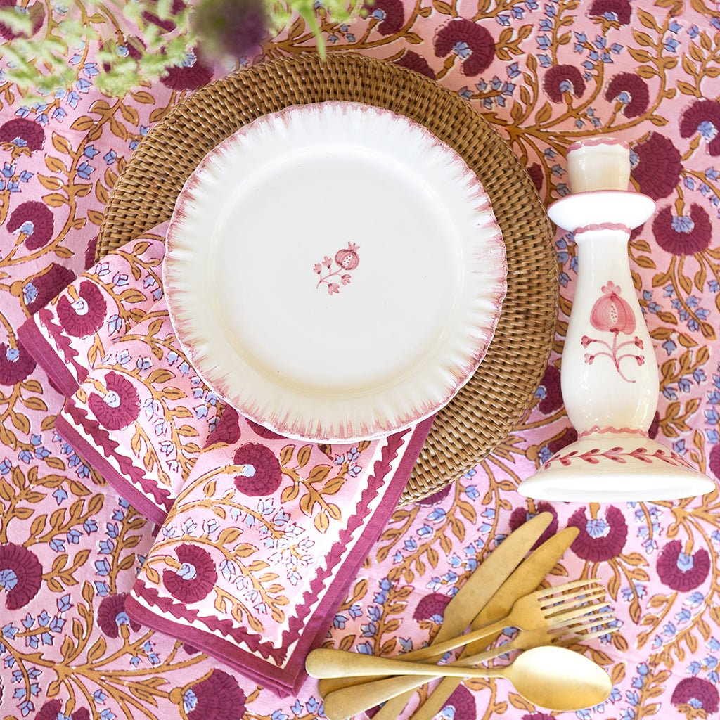 Table setting with a pink floral tablecloth, white plates, gold cutlery, and a decorative bottle.