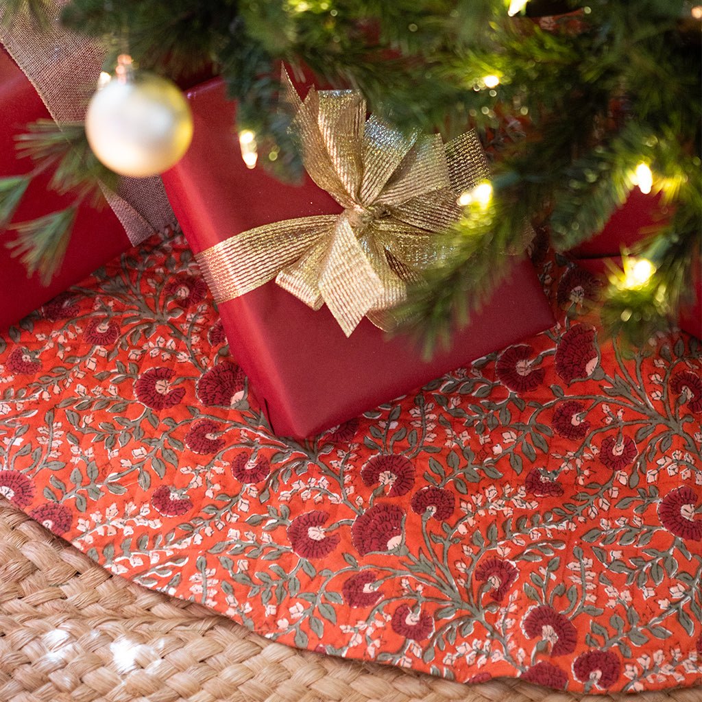 Red gift box with gold bow under a Christmas tree on a decorative tablecloth.