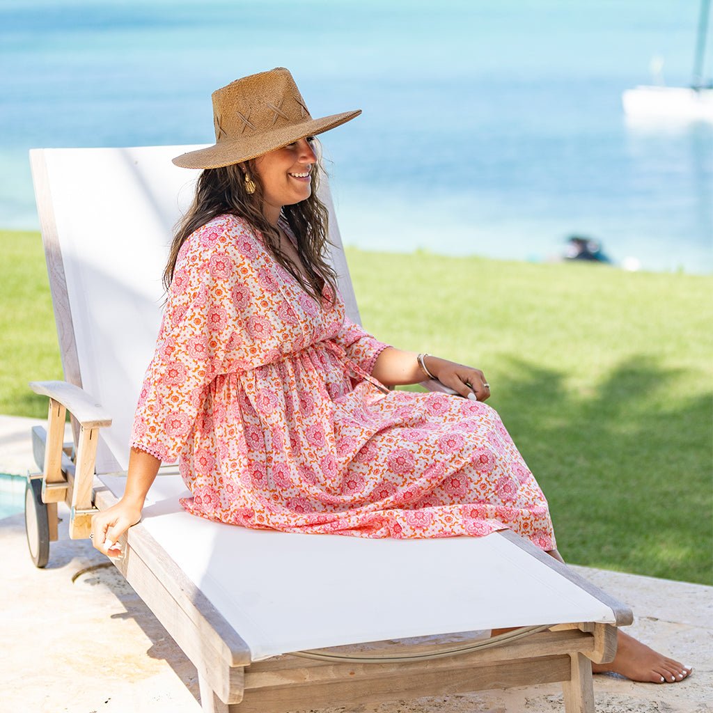 Woman in a floral dress and straw hat sitting on a lounge chair by the water.