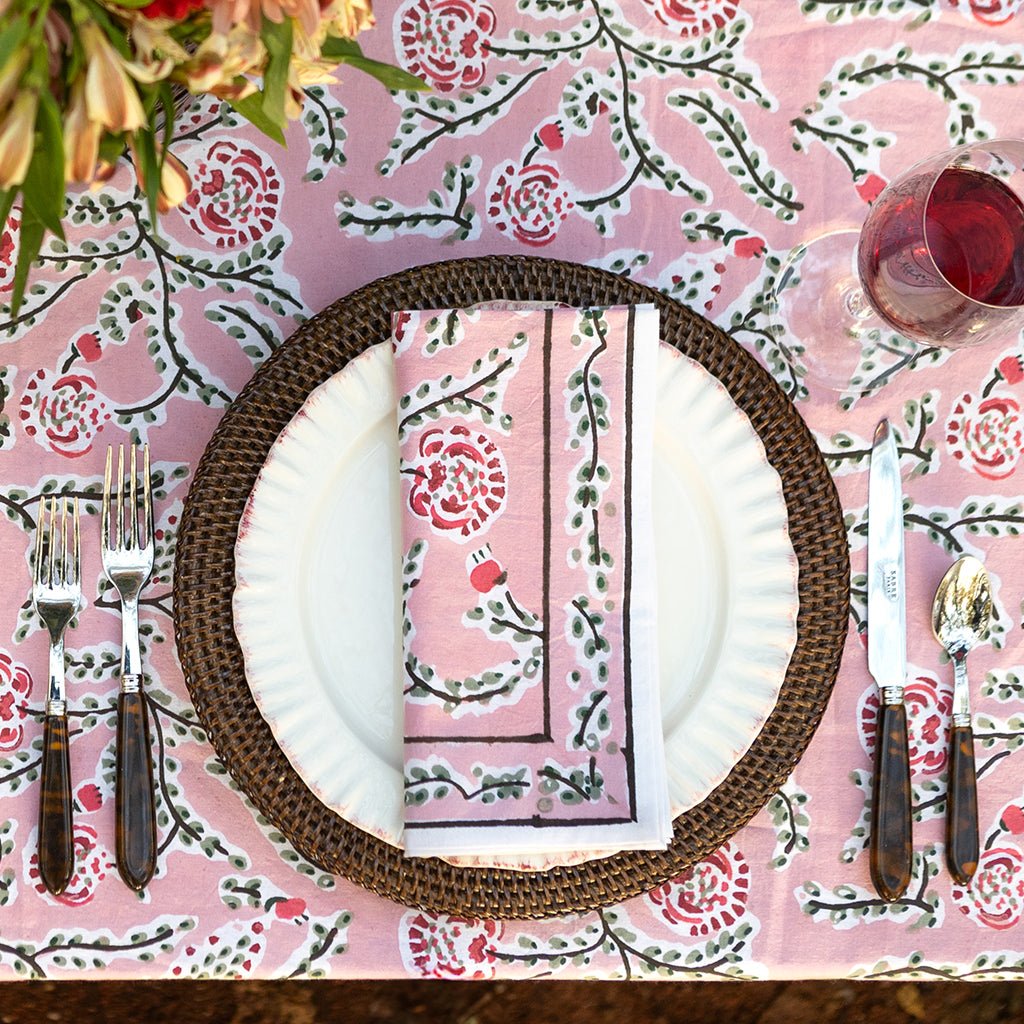 Table setting with floral tablecloth, plates, cutlery, and napkin on a decorative pink and green tablecloth.
