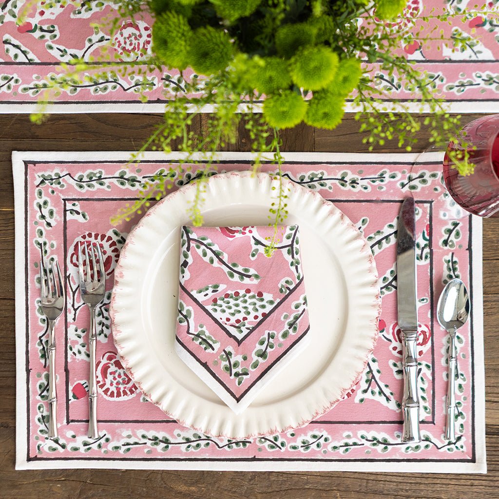 Table setting with a pink decorative placemat, white plate, and silverware on a wooden table.