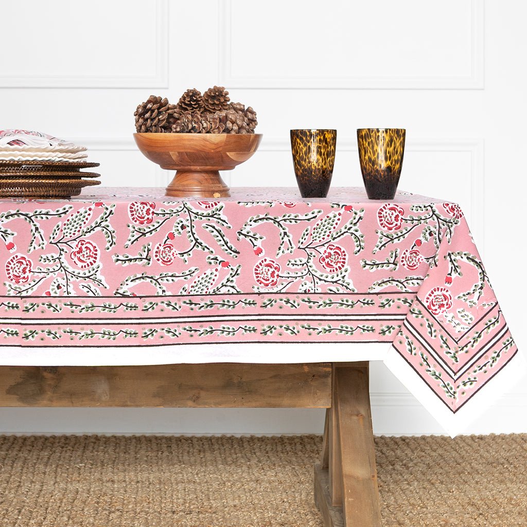 Table with a pink floral tablecloth, wooden bowl, and glasses on a white wall background.