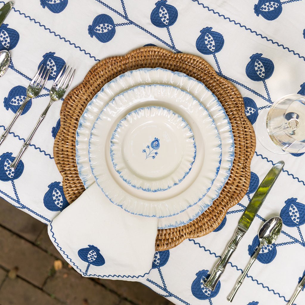 Table setting with woven placemat, white plates, and blue patterned tablecloth.