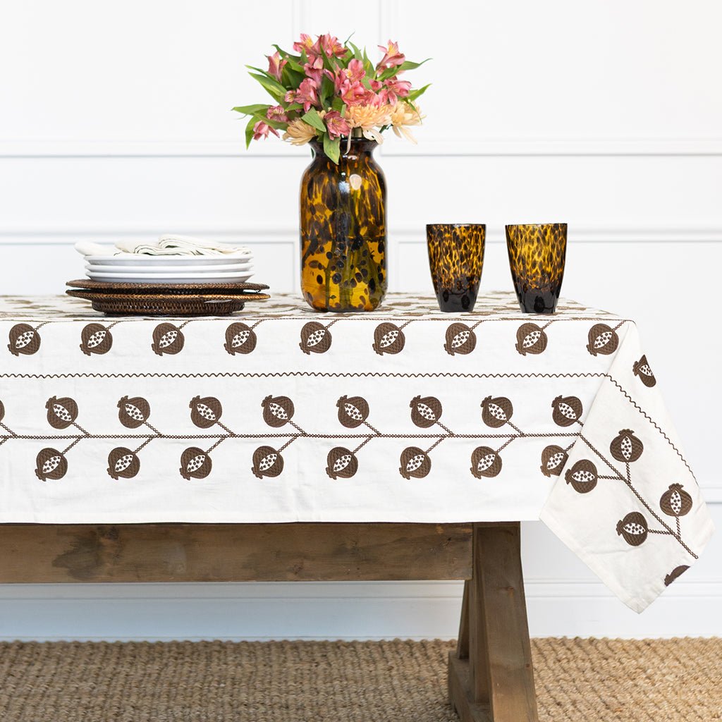 Table setting with a patterned tablecloth, vase with flowers, and glasses on a wooden table.