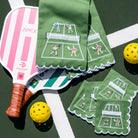Pickleball paddle with pink and white design, green tea towels with pickleball court illustration, and yellow balls on a green surface.