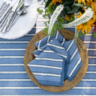 Blue and white striped napkin on a woven plate with a striped tablecloth and flowers in the background
