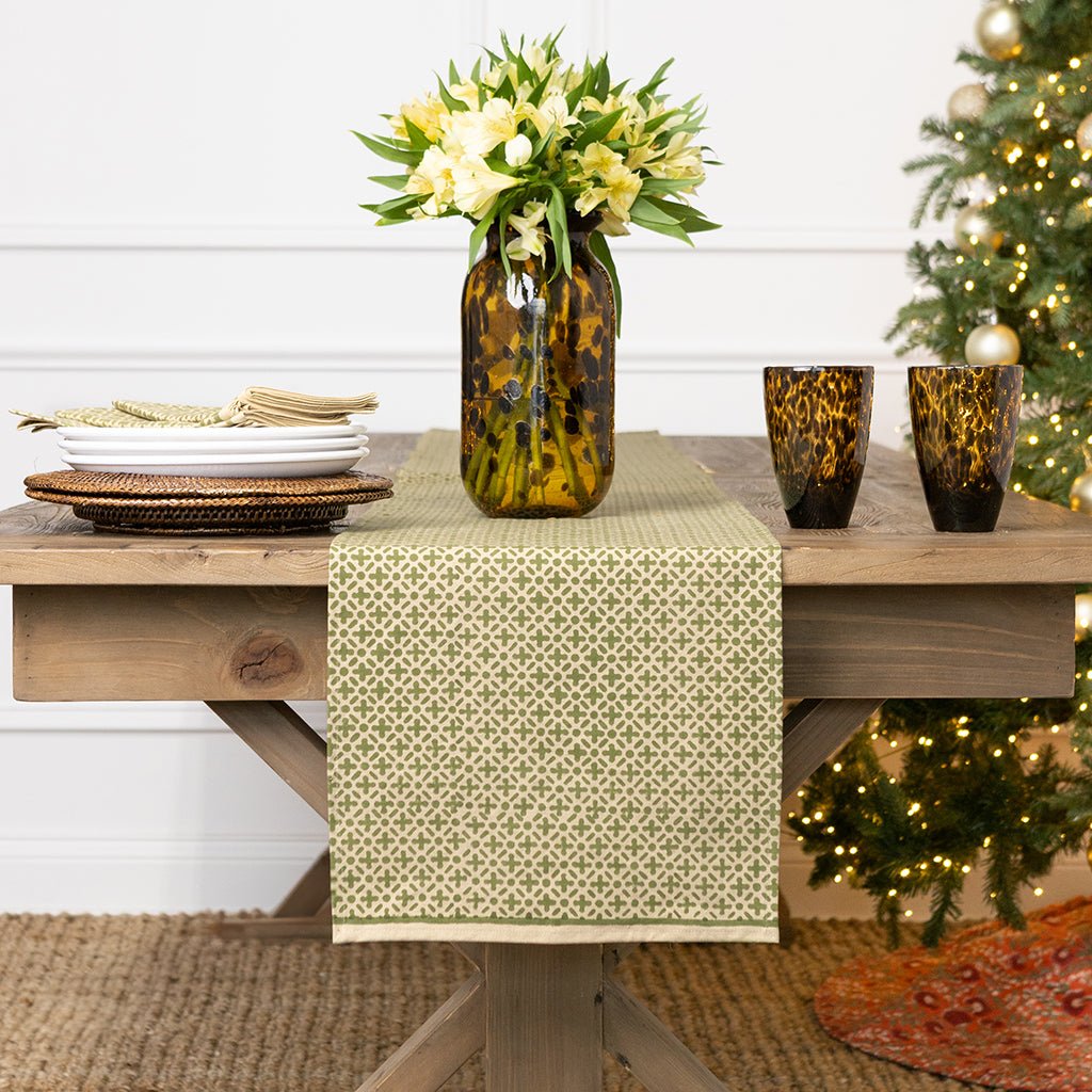 Table setting with a patterned table runner, vase with flowers, and decorative items against a Christmas tree.