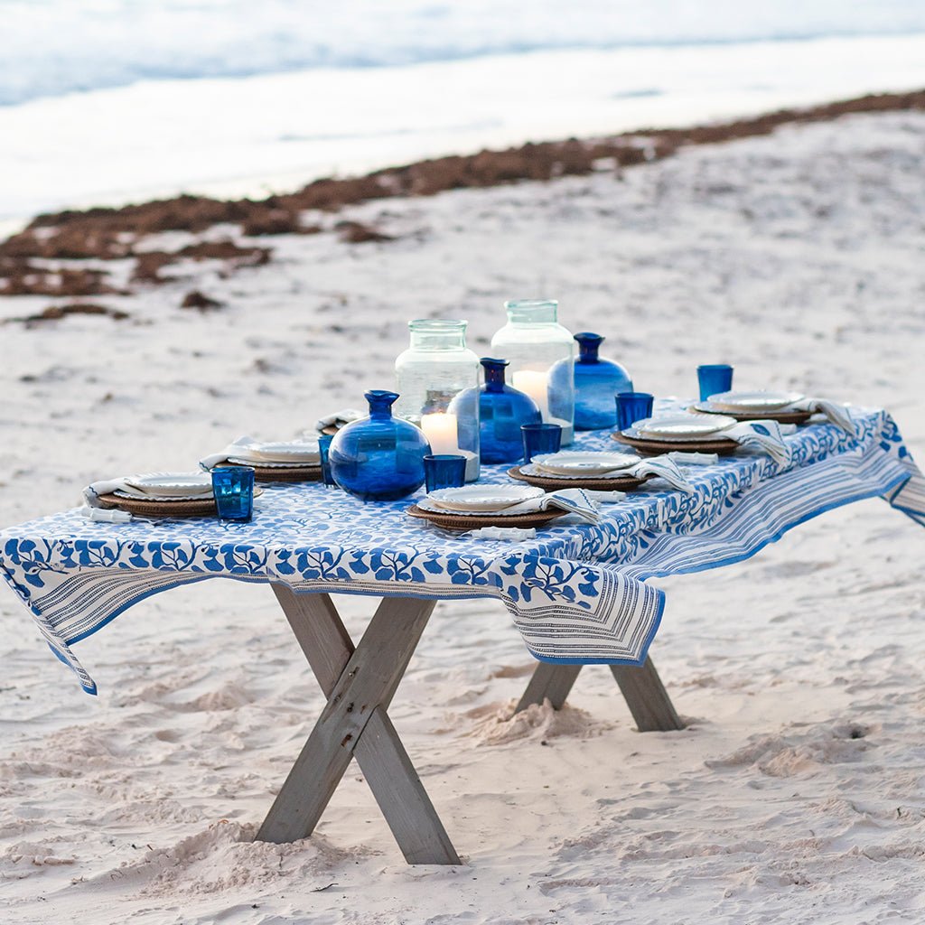 Dining table set with blue and white tablecloth, plates, and bottles on a sandy beach.