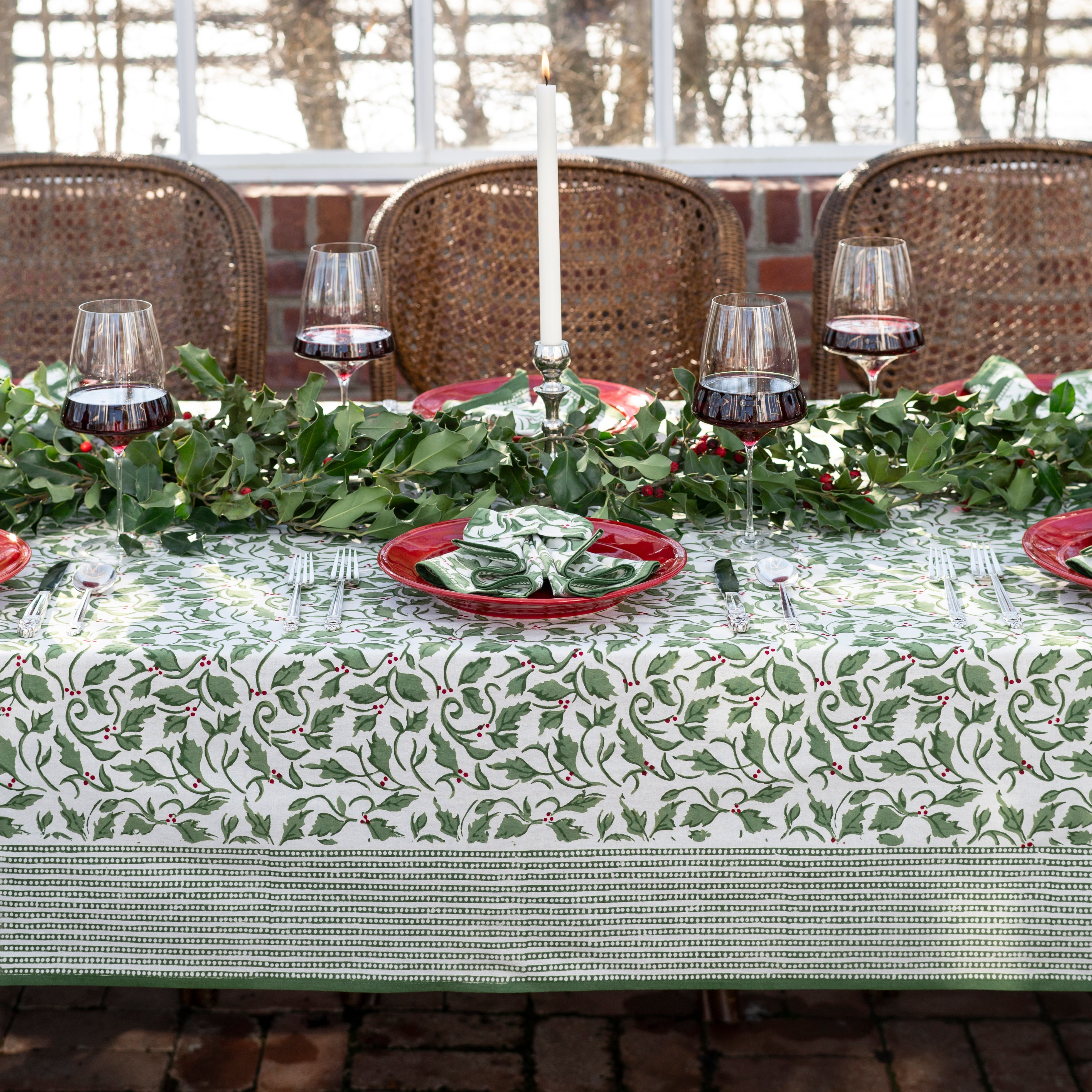 A set table covered with a green and white tablecloth featuring a holly berry pattern, arranged with dishes and silverware.
