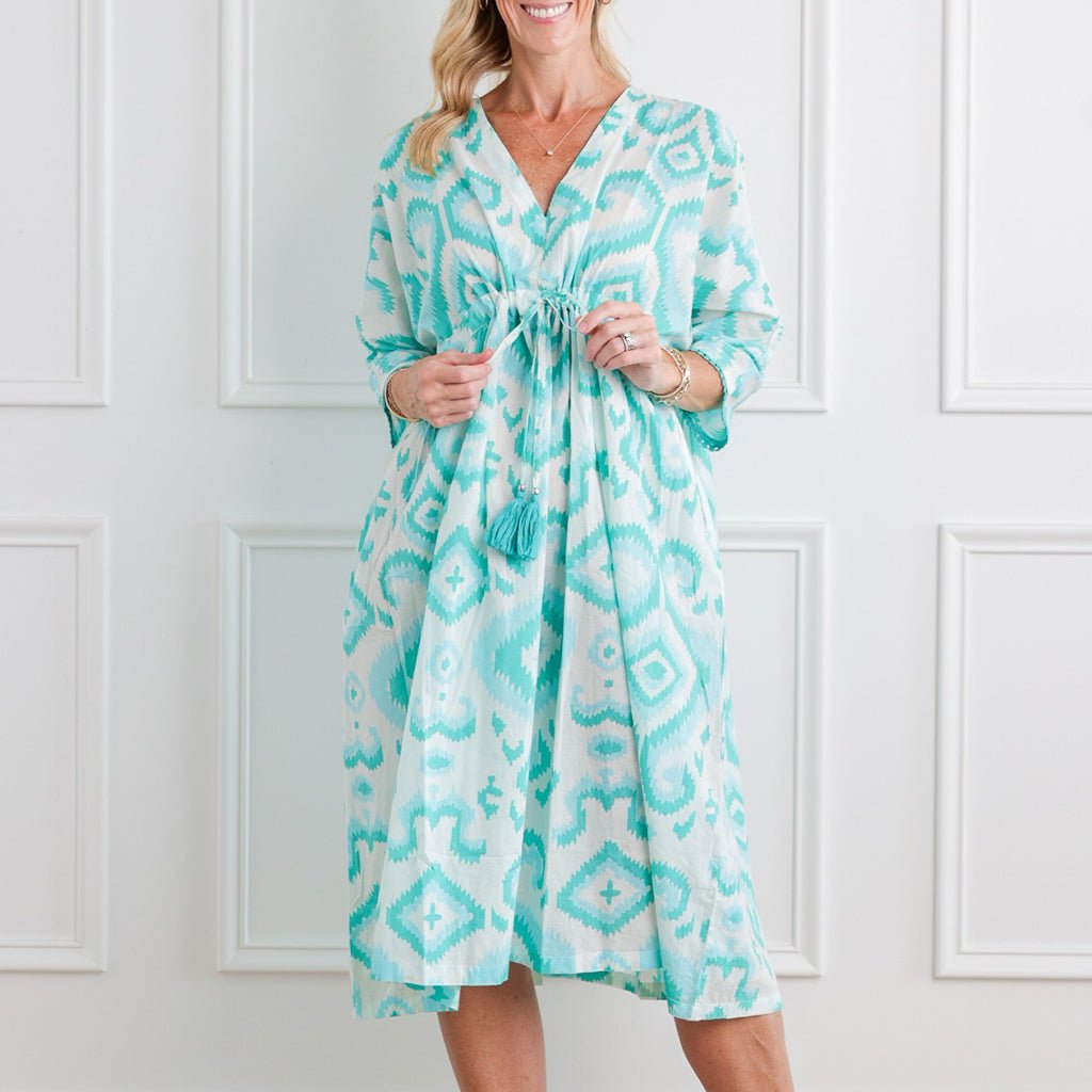 Woman wearing a turquoise patterned dress against a white paneled wall.