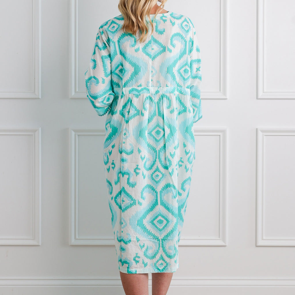 Person wearing a turquoise and white patterned dress against a white paneled wall.