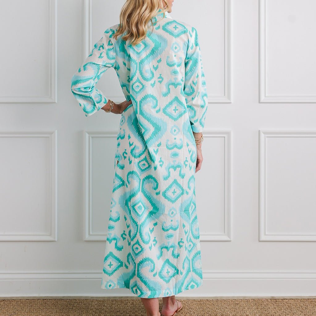 Woman wearing a long, patterned dress with turquoise and white design against a white paneled wall.