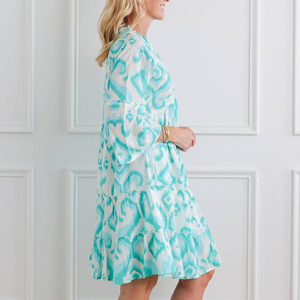 Woman wearing a turquoise and white patterned dress against a white paneled wall.