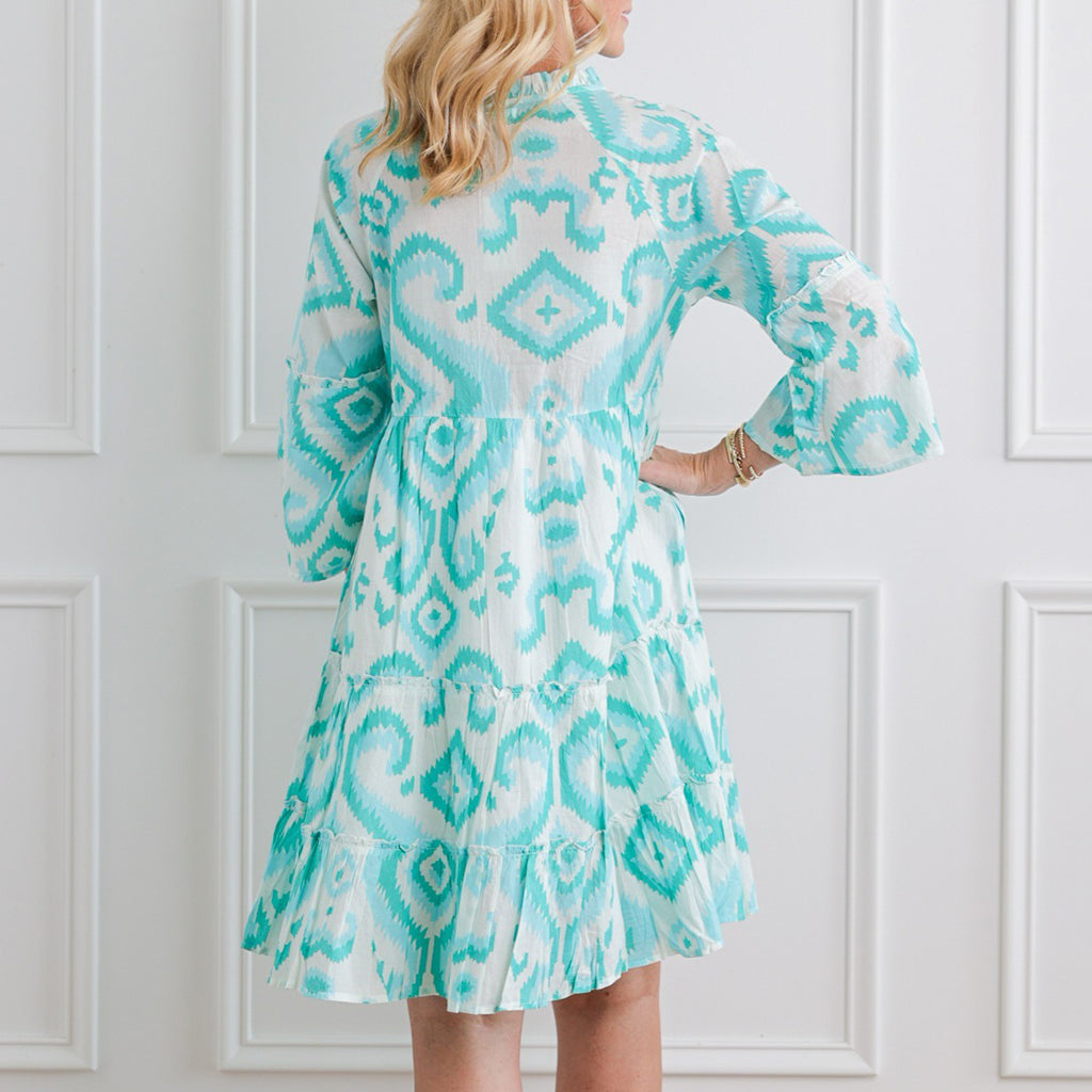 Woman wearing a turquoise patterned dress against a white paneled wall