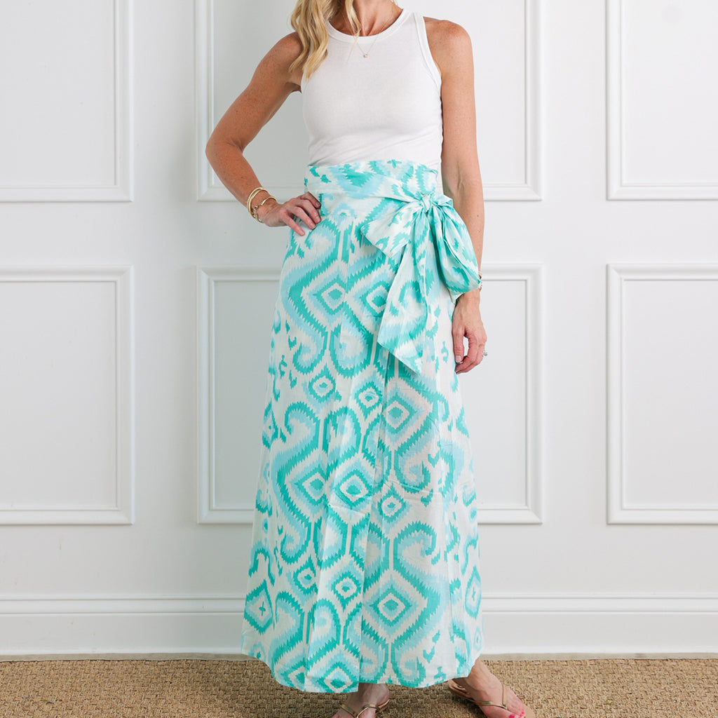 Woman wearing a white sleeveless top and a long turquoise patterned skirt against a white paneled wall.