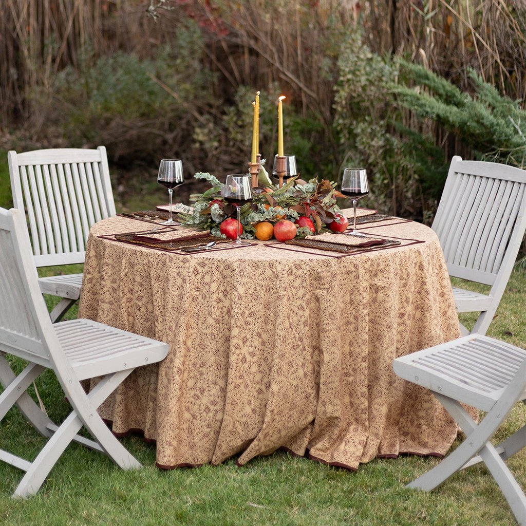 Decorated outdoor table with patterned tablecloth, candles, and fruit surrounded by white chairs.