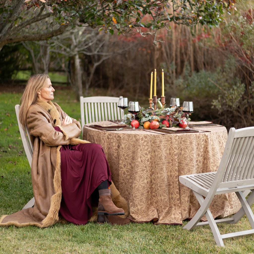 Woman in a fur coat sitting at an outdoor table set for a meal with candles and fruit.