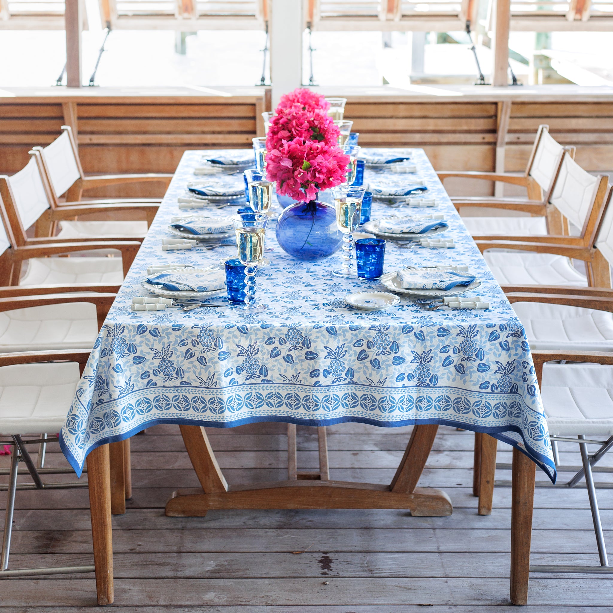 Dining table set with a blue floral tablecloth, pink flowers, and blue glasses on a wooden deck.