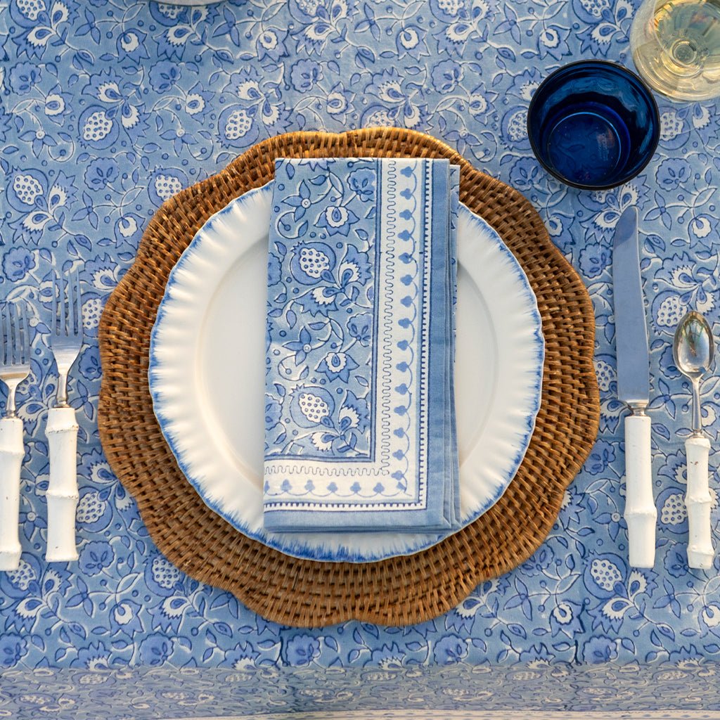 Table setting with blue and white patterned tablecloth, brown charger plate, and white plates with blue napkins.