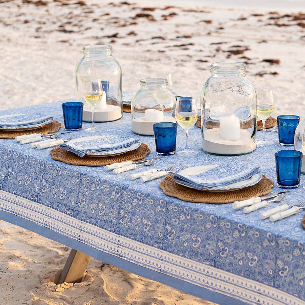 Dining table set with blue tablecloth, plates, glasses, and candles on a sandy beach.