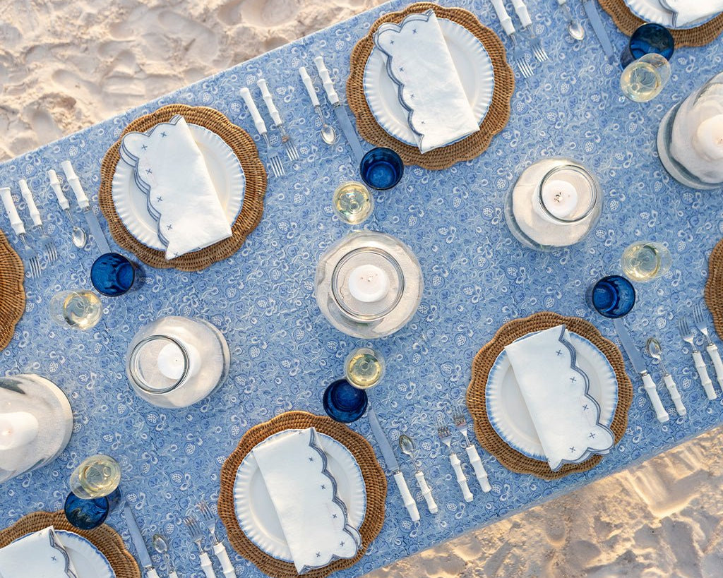 Dinner table setting with blue runner, white plates, and candles on a sandy surface.