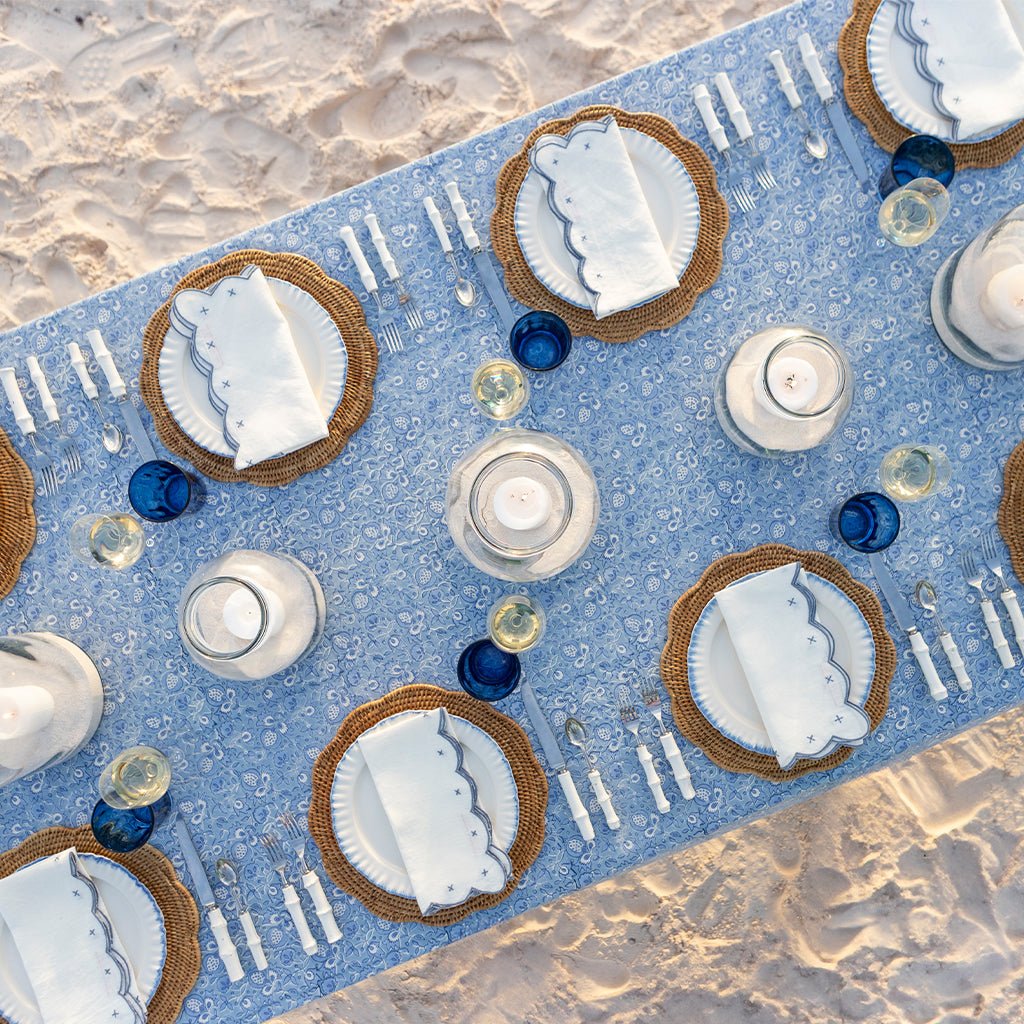 Dinner table setting with blue runner, white plates, and candles on a sandy surface.