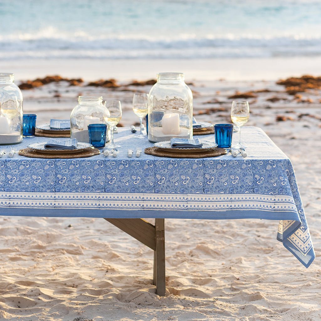 Dining table set with blue and white tablecloth, glasses, and candles on a sandy beach.