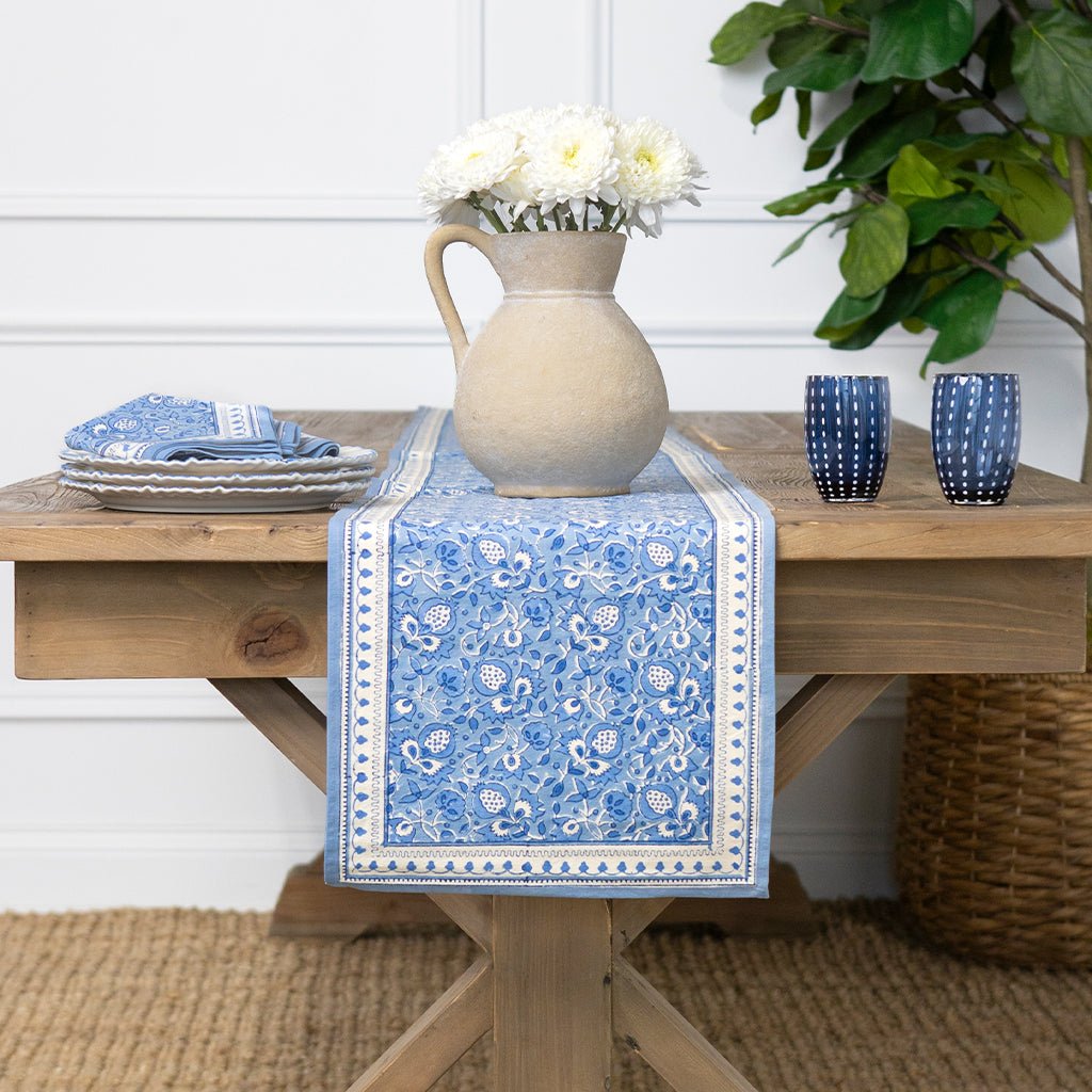 Wooden table with a blue patterned table runner, white flowers in a vase, and blue cups.