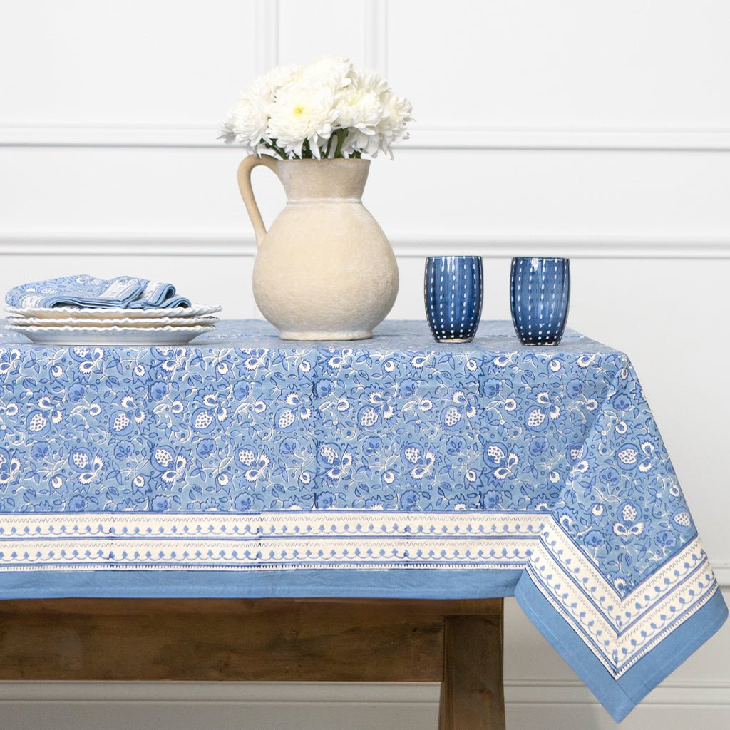 Blue patterned tablecloth on a wooden table with white flowers and blue glasses.