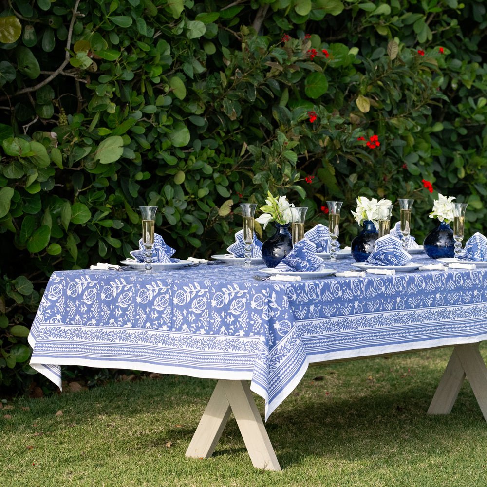 A blue and white floral patterned tablecloth is displayed on an outdoor table set for dining, with lush greenery in the background.