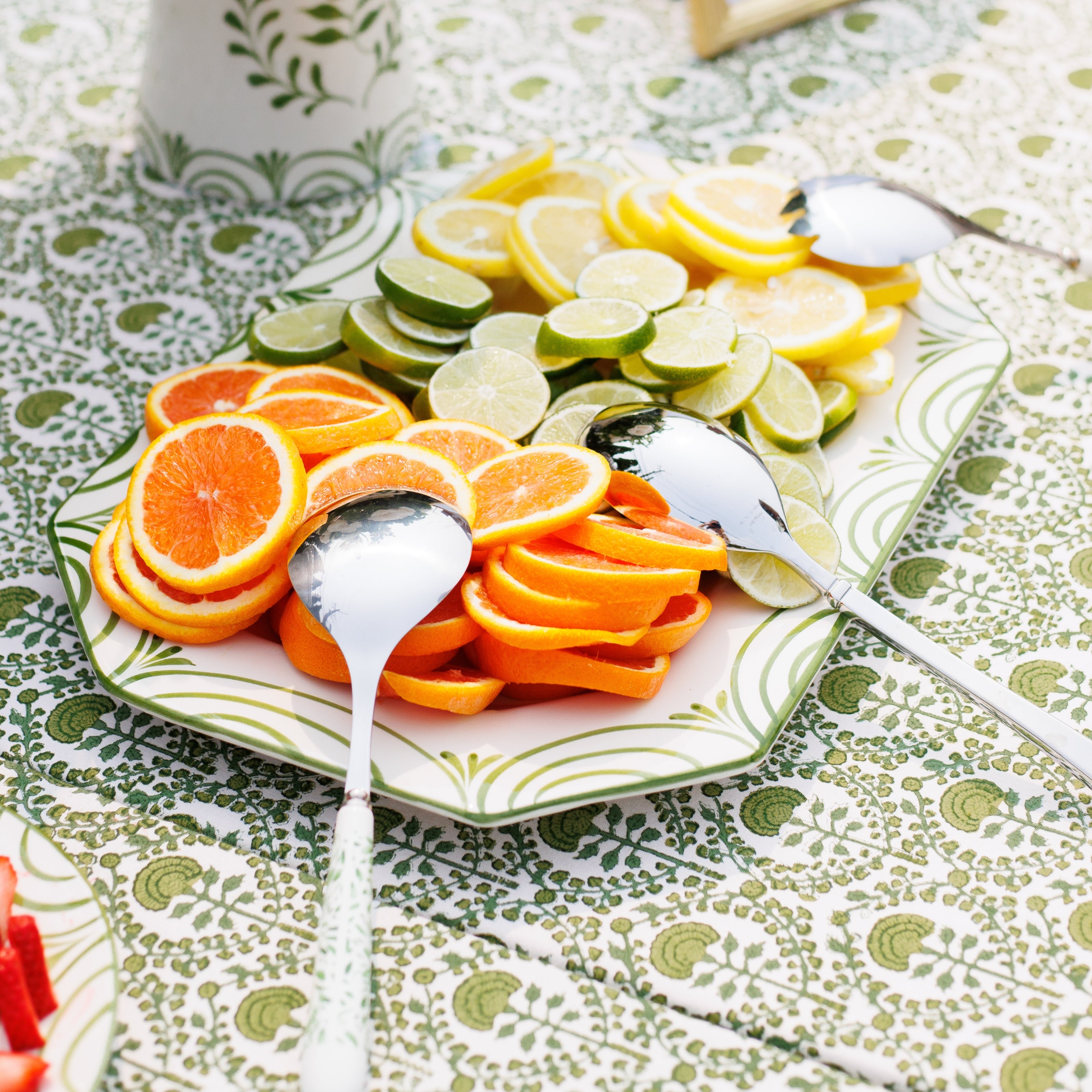 Sliced oranges and limes on a platter with spoons on a patterned tablecloth.