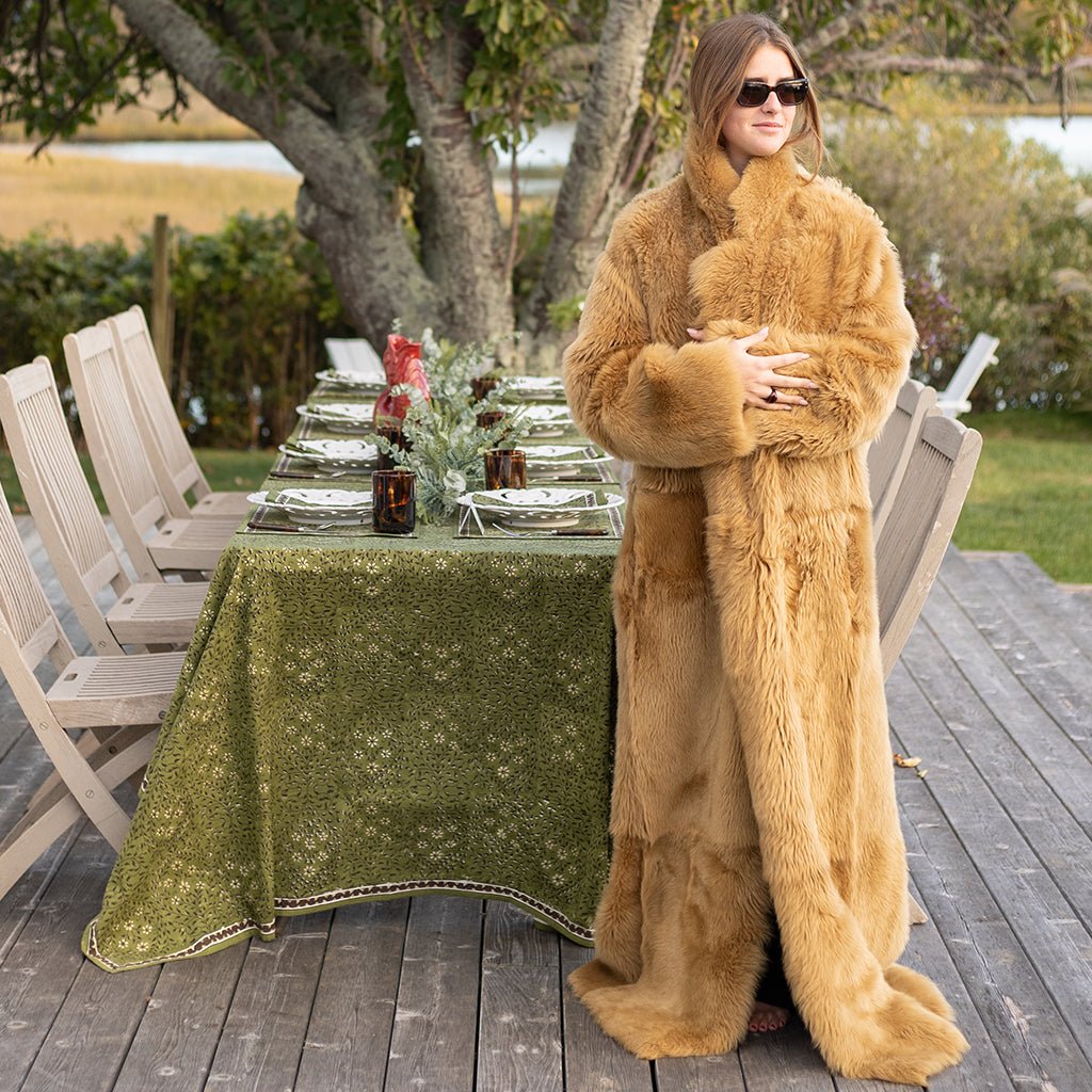 Woman in a fur coat standing by an outdoor dining table with a green tablecloth.