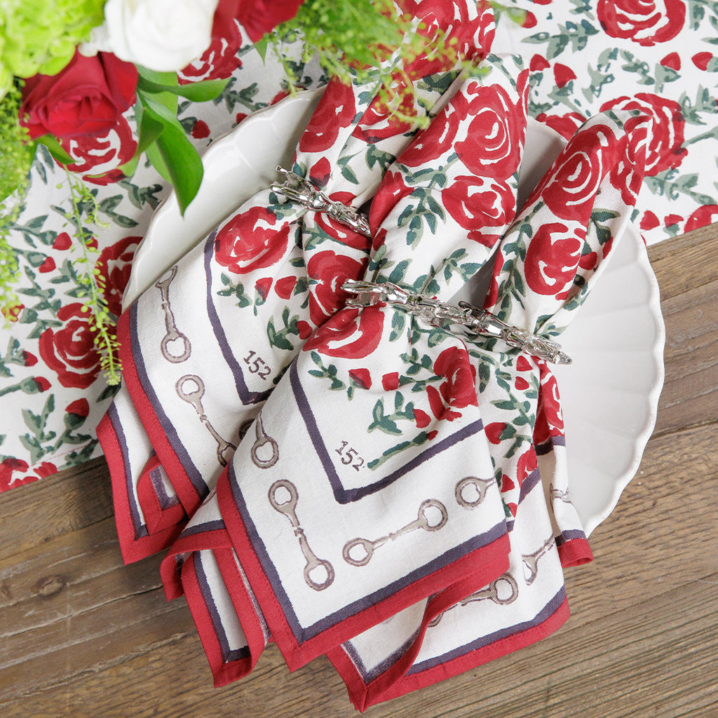 Set of four red rose patterned napkins with a snaffle bit border on a wood table.