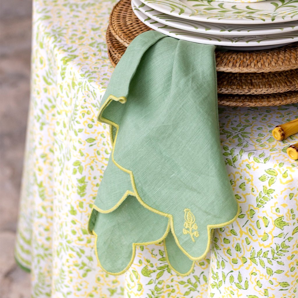 Green embroidered napkin hanging on rattan placemats with plates underneath on a patterned tablecloth.