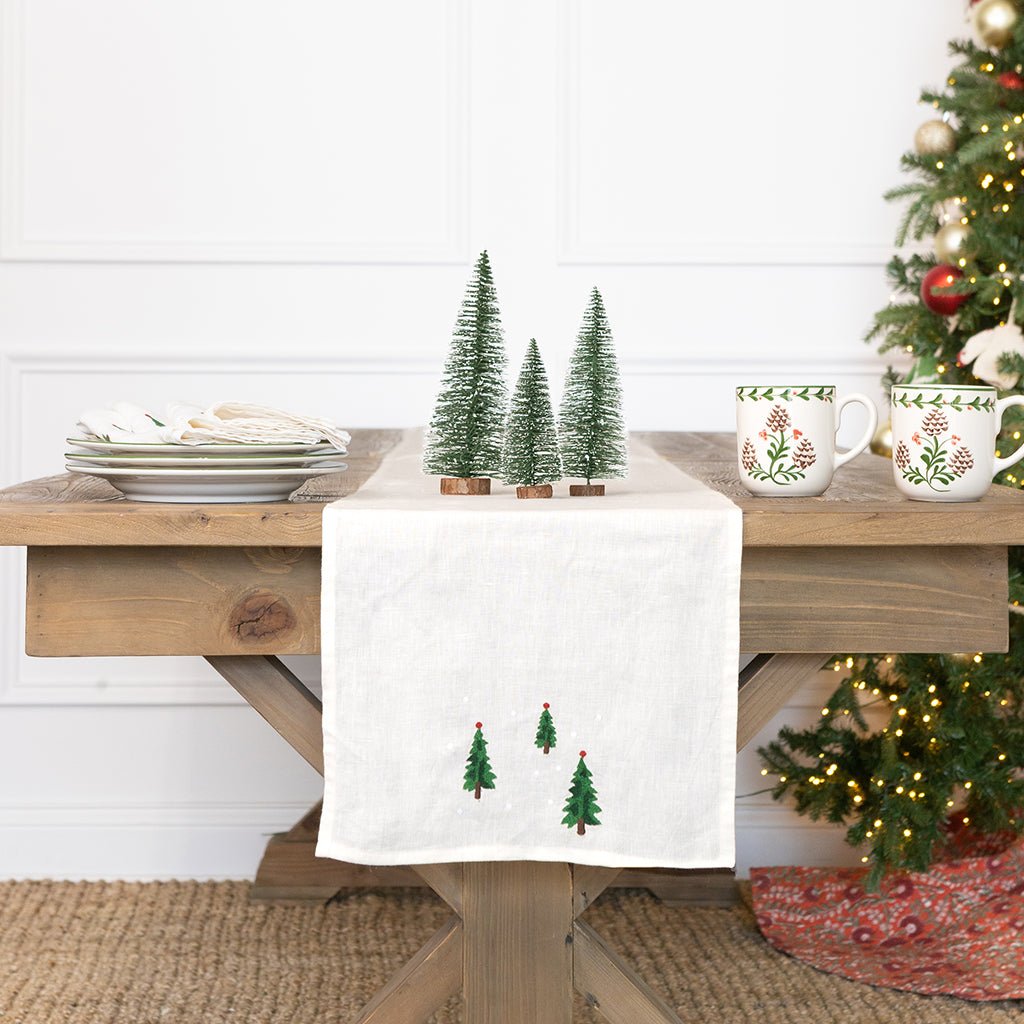 Wooden table with Christmas decor including a white towel with tree designs, mugs, and a decorated Christmas tree.