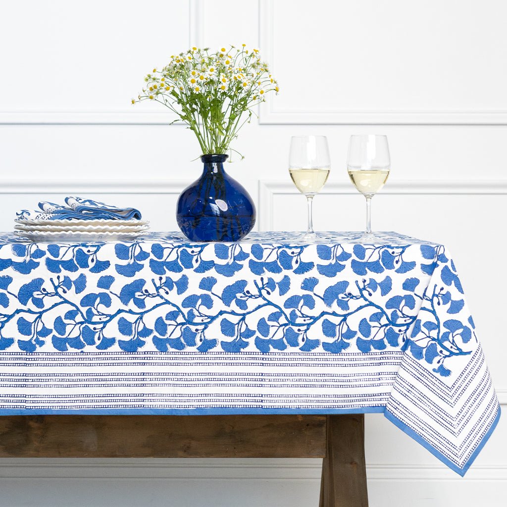 Table with a blue and white floral tablecloth, vase, and glasses on a white wall background.