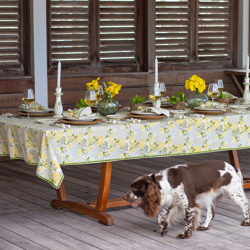 Dog standing next to a table set for a meal with floral tablecloth and yellow flowers.