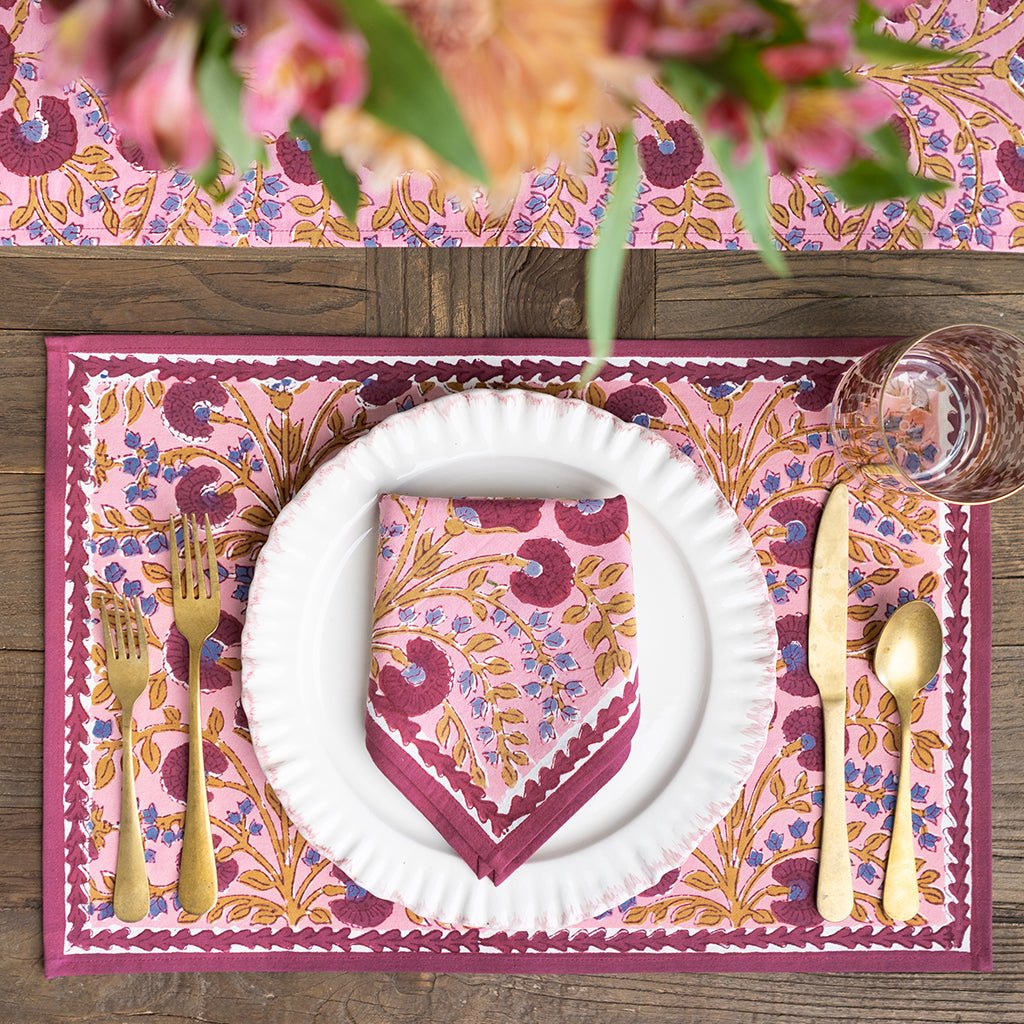 Dining table setting with pink floral placemat, plate, and cutlery on a wooden surface.