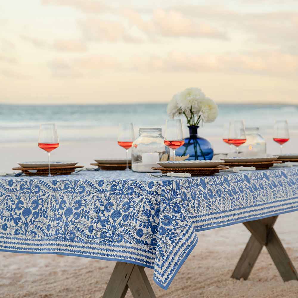 A cornflower blue and white floral designed tablecloth laid out on a table with a glass of red wine and dinnerware in the background, set against a beach and ocean backdrop.