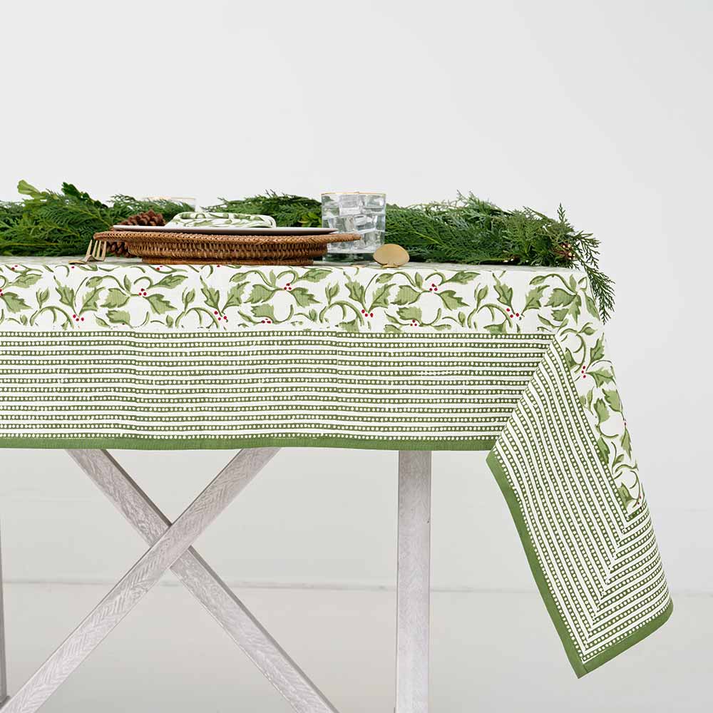 A set table covered with a green and white tablecloth featuring a holly berry pattern, arranged with dishes and silverware.