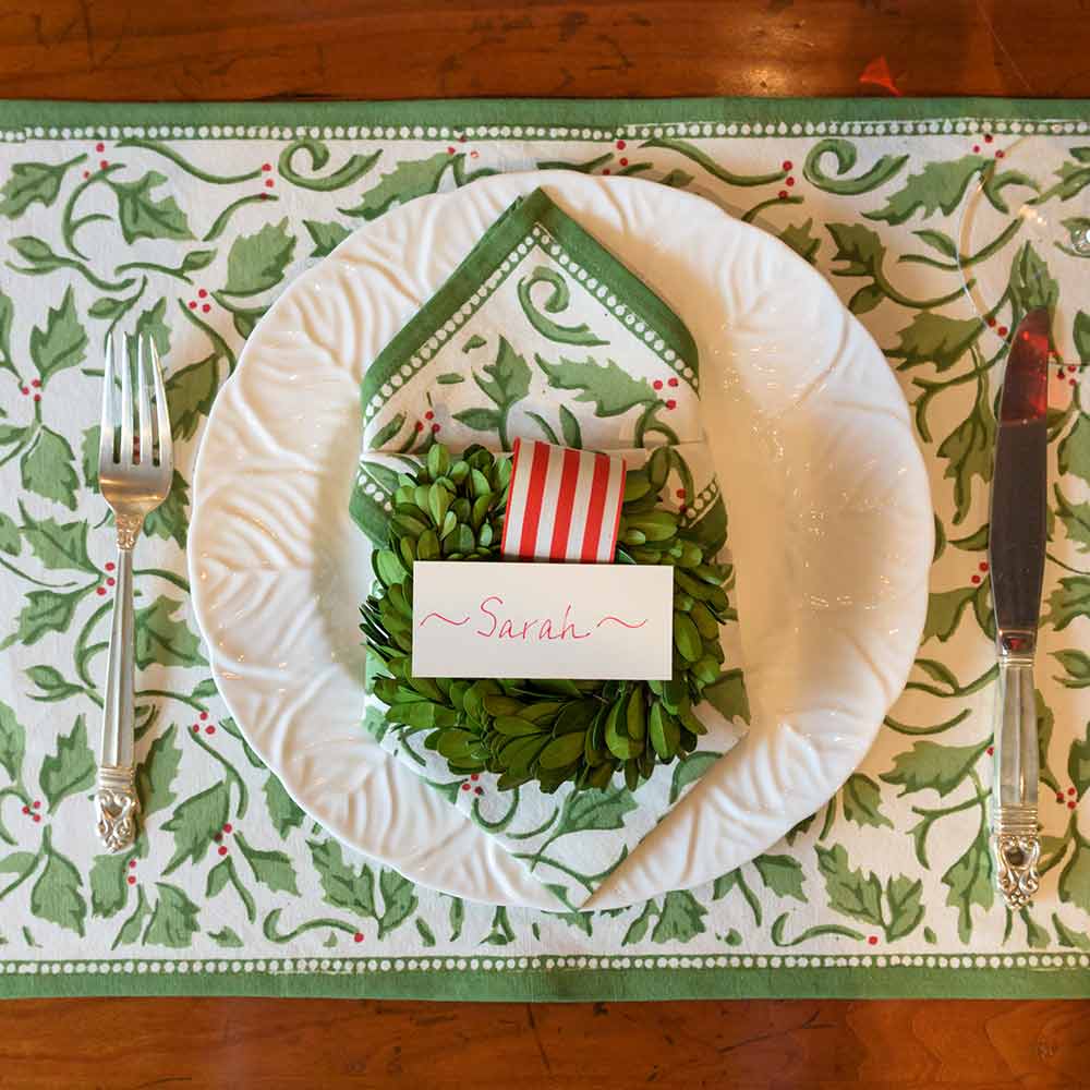 A set of four cotton placemats with a Christmas holly and berries pattern displayed on a table setting alongside a plate and cutlery.