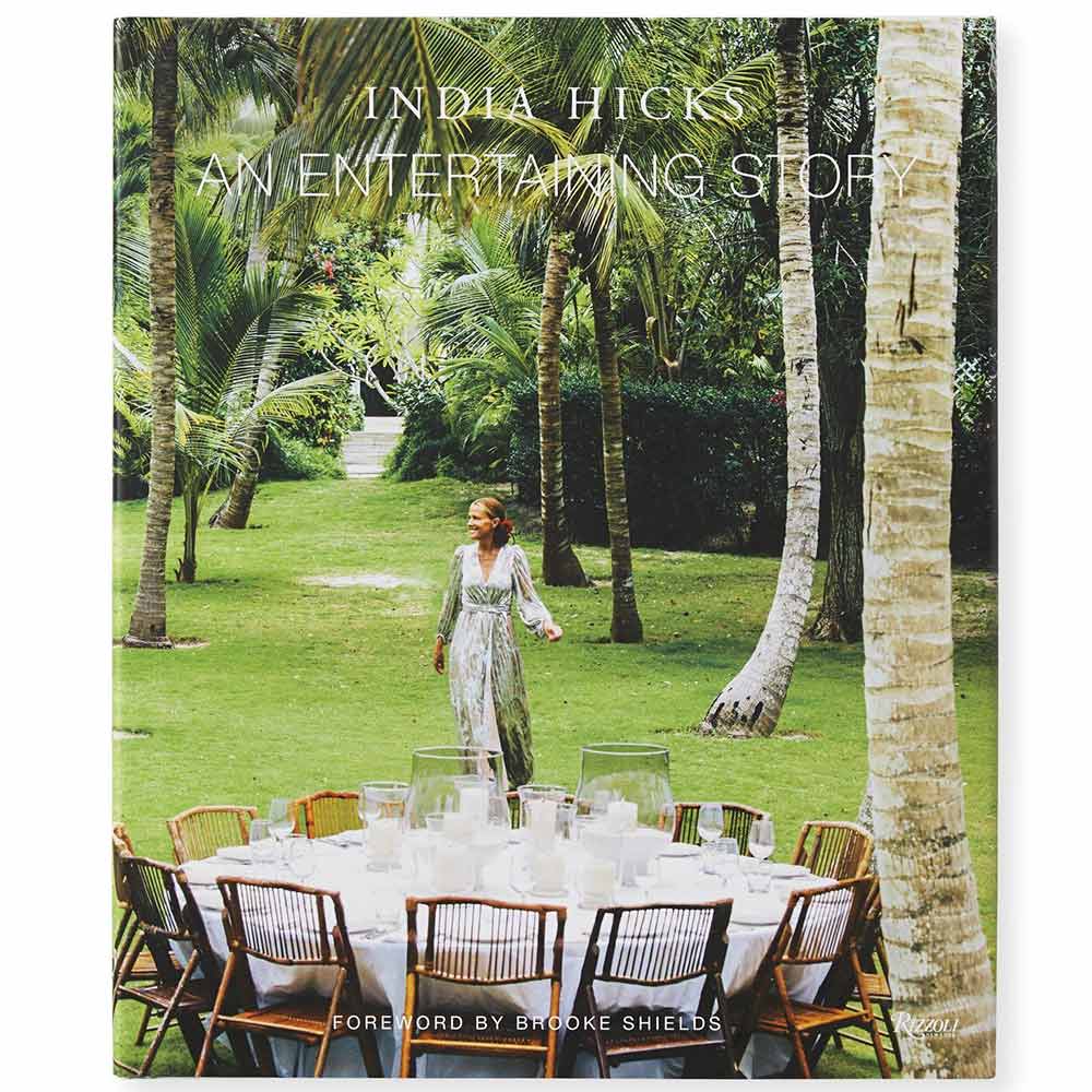 A cover image of a book titled 'An Entertaining Story' by India Hicks, featuring a woman standing by a table laid out with glasses and a pitcher, in an outdoor setting with palm trees in the background.
