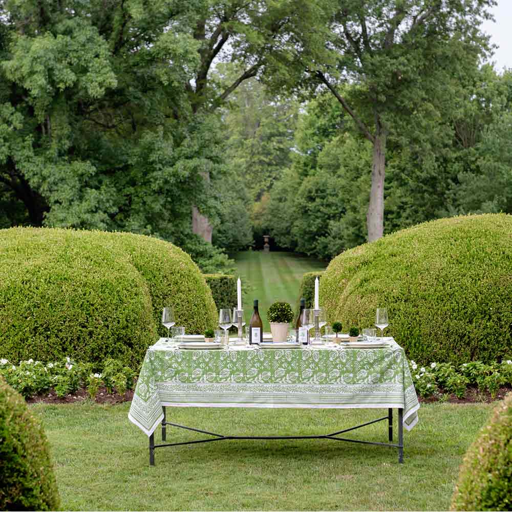 Outdoor table with Pomegranate Green tablecloth surrounded by green trees and bushes. 