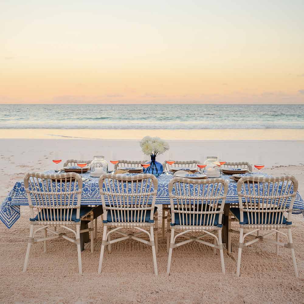 Dining table set for four with a blue tablecloth on a sandy beach at sunset.